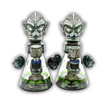 Artistic glass dab rig with tribal warrior mask topper, engraved stone-style accents, and clear beaker chamber with decorative side handles.