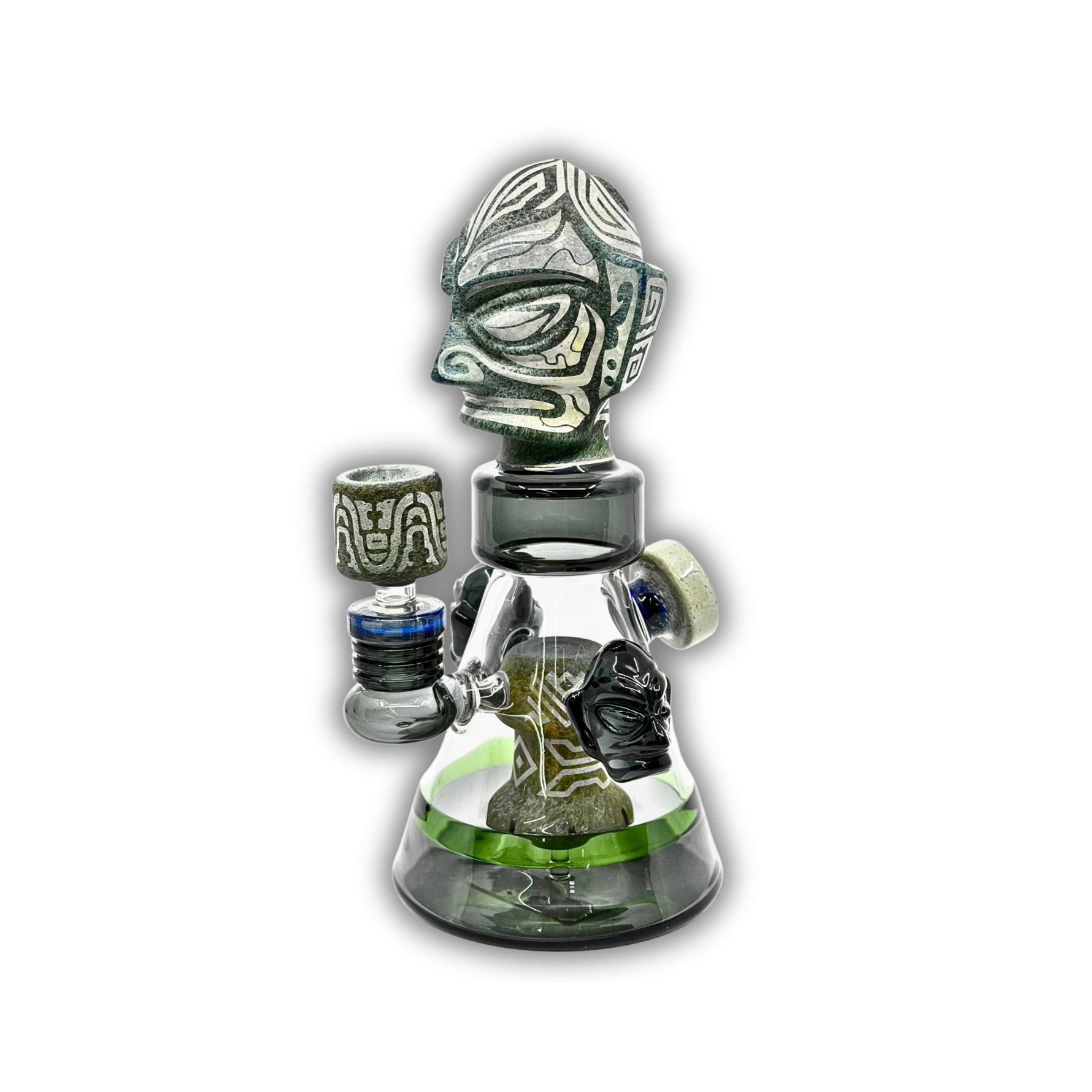 Artistic glass dab rig with tribal warrior mask topper, engraved stone-style accents, and clear beaker chamber with decorative side handles.