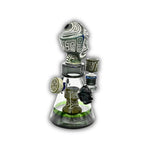 Artistic glass dab rig with tribal warrior mask topper, engraved stone-style accents, and clear beaker chamber with decorative side handles.